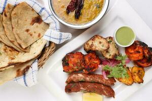 Mixed grill kebab chicken mutton beef prawn fish mint sauce lemon pickled onion bread basket chapati naan tandoori paratha dal lentil fry tadka copper pot on white dish over white background photo