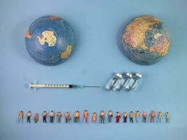 Miniature human figure figurine male female doll row line vaccine bottle medical injection syringe needle world map globe border copy text sign space on blue paper background photo