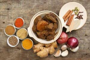 Beef bhuna dry curry with potato in ceramic bowl ingredients chilli cumin coriander turmeric powered salt onion garlic ginger bay leaf cinnamon cardamom clove over rustic wood background photo