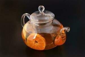 Liquid tea lemon orange slice green leaf cinnamon stick in transparent glass teapot kettle on black background photo