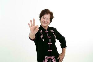 Elderly asian Chinese female wearing black tang samfu on white background happy smile ok finger sign at camera photo