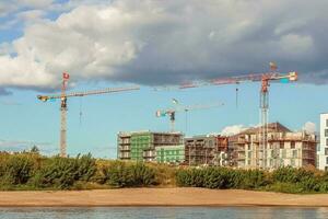 Construction site with three cranes with three flags Ukrainian, Lithuanian and Lithuanian coat of arms building residential buildings on river bank with sandy beach photo