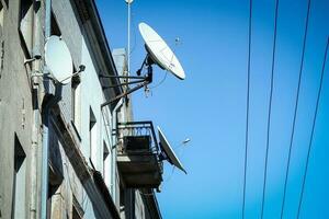 White satellite dish antennas on the roof and wall of ann old building with lines or wires on blue sky background photo