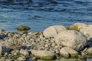 Close up of stacks of rocks on the river bank near dark blue wavy water stream on sunny day photo