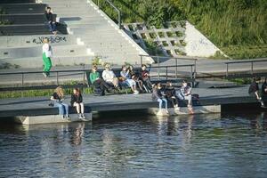 Vilna, Lituania 03 06 2022 joven personas y personas mayores sentado cerca Neirs río Relajado en sepia calentar puesta de sol ligero foto