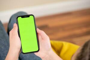 joven niña sentado en un sofá y utilizando un teléfono inteligente con verde pantalla foto