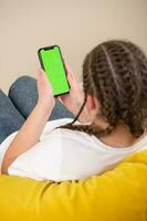 Young girl sitting on a sofa and using a smartphone with green screen photo