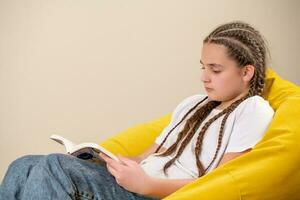 Teenage girl with braids reading book on yellow beanbag chair photo