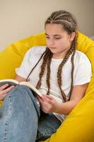 Teenage girl with braids reading book on yellow beanbag chair photo