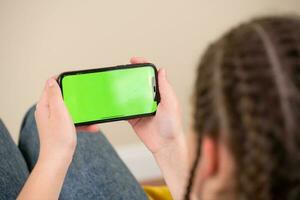 joven niña sentado en un sofá y utilizando un teléfono inteligente con verde pantalla foto