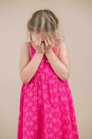 pequeño niña en un rosado vestir en un beige antecedentes cubre su cara con su manos foto