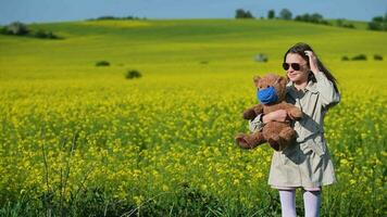 Jeune caucasien fille permanent dans milieu de colza Prairie en portant et pressant jouet nounours ours portant bleu médical visage masque. video