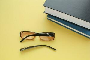 Old broken eyeglasses with damaged legs and books on yellow background. Poor eyesight. Repair concept. Idea of health. Failure optic eyewear. Breakage of vision correction glasses. Close up, flat lay photo