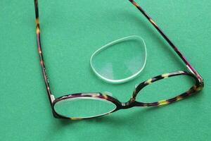 Old broken eyeglasses with damaged lens on green background. Poor eyesight. Reuse and repair concept. Idea of health. Failure optic eyewear. Breakage of vision correction glasses. Close up, flat lay photo