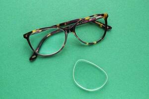 Old broken eyeglasses with damaged lens on green background. Poor eyesight. Reuse and repair concept. Idea of health. Failure optic eyewear. Breakage of vision correction glasses. Close up, flat lay photo