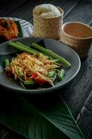 Thai food dish both in Thailand and Asia, Papaya Salad or as we call it Somtum is complemented with grilled chicken and sticky rice with fresh stir-fries. Served on the black wooden table. photo