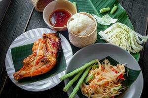 Thai food dish both in Thailand and Asia Papaya Salad or as we call it Somtum is complemented with grilled chicken and sticky rice with fresh stir-fries. Served on the black wooden table. photo