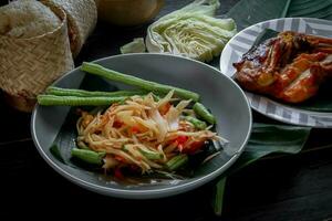 tailandés comida plato ambos en Tailandia y Asia papaya ensalada o como nosotros llamada eso somtum es complementado con A la parrilla pollo y pegajoso arroz con Fresco frituras. servido en el negro de madera mesa. foto