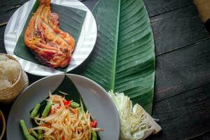 Thai food dish both in Thailand and Asia Papaya Salad or as we call it Somtum is complemented with grilled chicken and sticky rice with fresh stir-fries. Served on the black wooden table. photo