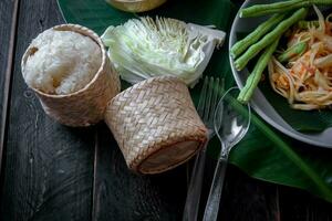 Thai food dish both in Thailand and Asia Papaya Salad or as we call it Somtum is complemented with grilled chicken and sticky rice with fresh stir-fries. Served on the black wooden table. photo