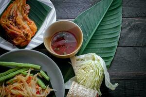 Thai food dish both in Thailand and Asia Papaya Salad or as we call it Somtum is complemented with grilled chicken and sticky rice with fresh stir-fries. Served on the black wooden table. photo