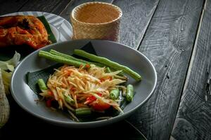 Thai food dish both in Thailand and Asia, Papaya Salad or as we call it Somtum is complemented with grilled chicken and sticky rice with fresh stir-fries. Served on the black wooden table. photo