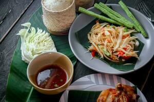 Thai food dish both in Thailand and Asia Papaya Salad or as we call it Somtum is complemented with grilled chicken and sticky rice with fresh stir-fries. Served on the black wooden table. photo