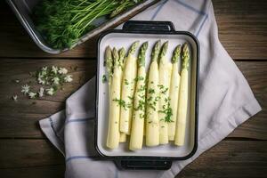 Traditional steamed white asparagus with hollandaise sauce and herbs as a top view in an enamel tray on an old wood table with copy space on the right, generate ai photo