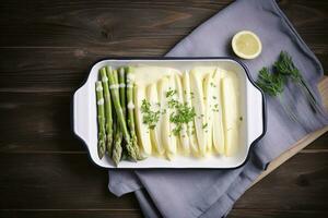 Traditional steamed white asparagus with hollandaise sauce and herbs as a top view in an enamel tray on an old wood table with copy space on the right, generate ai photo