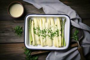 Traditional steamed white asparagus with hollandaise sauce and herbs as a top view in an enamel tray on an old wood table with copy space on the right, generate ai photo