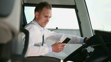 Masculin entraîneur chauffeur sur Pause séance à l'intérieur de cabine de garé autobus vérification mobile téléphone. video