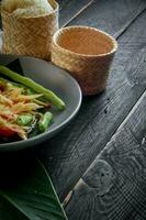 Thai food dish both in Thailand and Asia, Papaya Salad or as we call it Somtum is complemented with grilled chicken and sticky rice with fresh stir-fries. Served on the black wooden table. photo