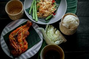 Thai food dish both in Thailand and Asia, Papaya Salad or as we call it Somtum is complemented with grilled chicken and sticky rice with fresh stir-fries. Served on the black wooden table. photo