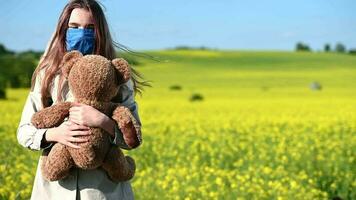 liten flicka med teddy Björn leksak bär skyddande ansikte mask stående i stor gul fält på jordbruksmark lekfullt spinning runt om. video