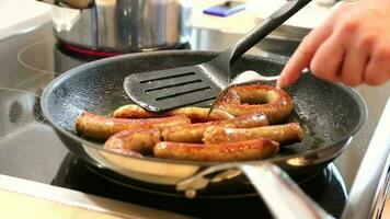 Woman cooking sausages in pan on kitchen cooking plate with fork and black spatula with delicious fatty bratwurst for dinner as unhealthy but spicy and roasted sausage meal for lunch and the children video
