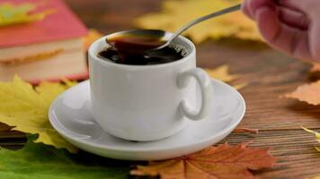 hand stirring coffe with spoon in cup on wooden table with book and colorful autumnal maple leaves video