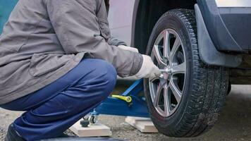 velho homem dentro sujo azul calça e cinzento Jaqueta removendo pneu parafusos Fora do prata carro video
