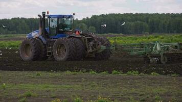Blue New Holland tractor with double wheels pulling disc harrow with roller basket at spring day with slow motion video