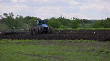 Blue New Holland tractor with double wheels pulling disc harrow with roller basket at spring day with slow motion video