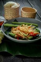 Thai food dish both in Thailand and Asia, Papaya Salad or as we call it Somtum is complemented with grilled chicken and sticky rice with fresh stir-fries. Served on the black wooden table. photo