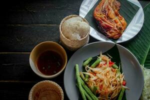 Thai food dish both in Thailand and Asia Papaya Salad or as we call it Somtum is complemented with grilled chicken and sticky rice with fresh stir-fries. Served on the black wooden table. photo