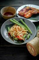 Thai food dish both in Thailand and Asia, Papaya Salad or as we call it Somtum is complemented with grilled chicken and sticky rice with fresh stir-fries. Served on the black wooden table. photo