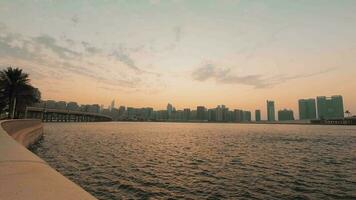 Zoom out Abu Dhabi city skyline on sunset time with water ripples timelapse from the breakwater near mall. Few clouds on orange sky hot evening video