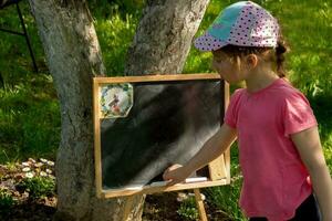 un niña en el jardín es yendo a dibujar en un pizarra tablero con tiza en un soleado verano día foto