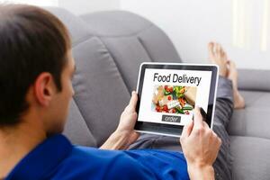 hombre ordenando tomar lejos comida por Internet con el tableta mientras acostado a hogar. foto
