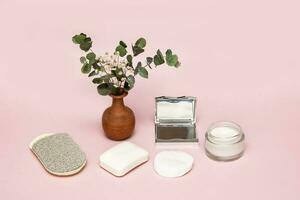 cosmetic accessories face sponge, soap, face cream, mirror, cotton pads on a soft pink background with a vase of greenery photo