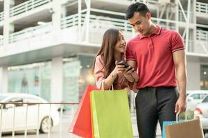 Happy young couple of shoppers walking in the shopping street towards and holding colorful shopping bags in hand and use a smartphone for check promotion. Concept of sale and Black Friday shopping photo