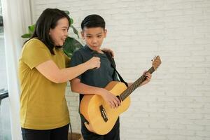 Asian boy playing guitar and mother sing a song and embrace, feel appreciated and encouraged. Concept of a happy family, learning and fun lifestyle, love family ties photo