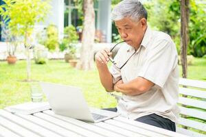 Portrait of old elderly Asian man using a computer laptop in the backyard for learning new skill after retired. Concept of no Ageism and not be late for learning. photo