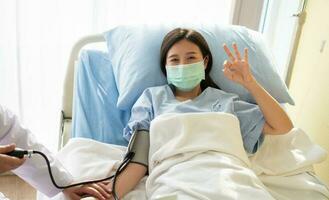 una paciente asiática feliz usa una máscara, se acuesta en la cama y muestra un símbolo de ok. cuando el médico examina los síntomas con un manómetro. concepto de creer en el tratamiento y la cobertura de seguro foto